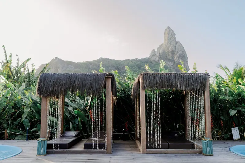 Dois bangalôs cobertos de palha no Dolphin Noronha, com o Morro do Pico ao fundo.