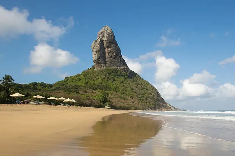 O Morro do Pico atrás de uma praia de areia dourada em Fernando de Noronha, sob céu azul.