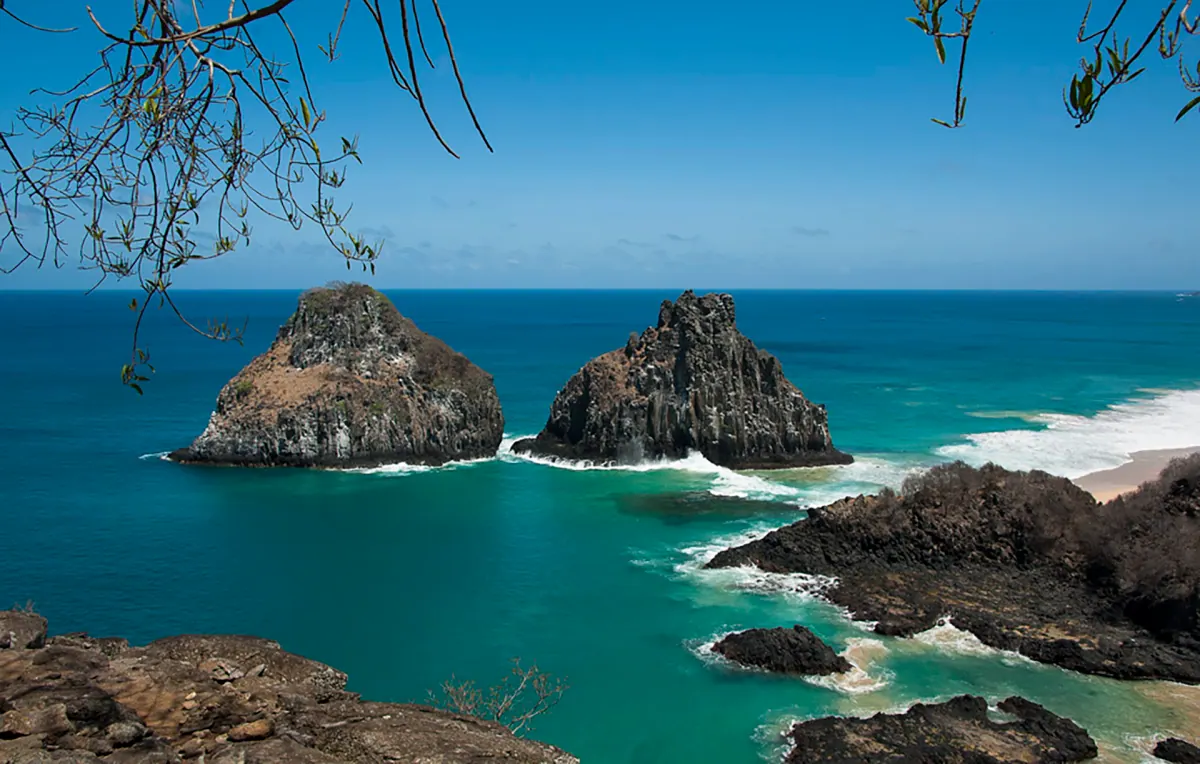 O Morro Dois Irmãos, dois morros rochosos no mar azul de Fernando de Noronha, visto entre galhos, com ondas quebrando nas pedras.