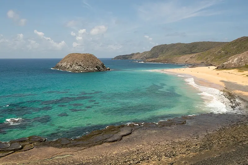 Praia de areia clara e mar verde em Fernando de Noronha, com uma ilhota rochosa perto da costa.
