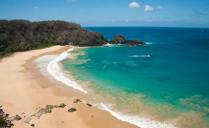 Praia extensa de areia clara e mar azul em Fernando de Noronha, vista do alto.