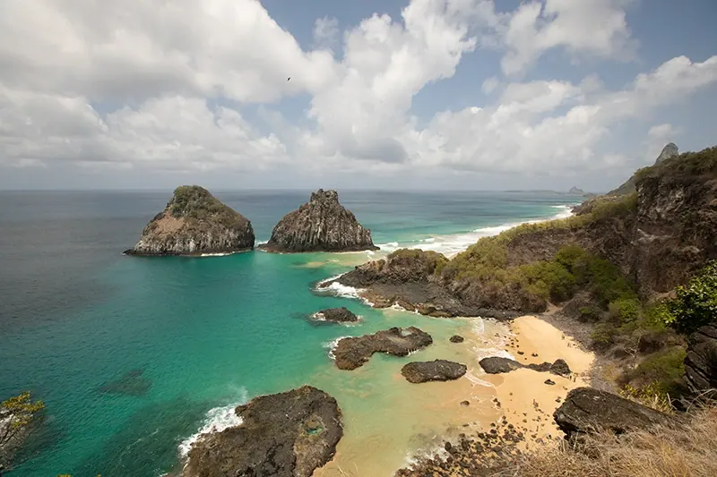 Vista aérea da costa de Fernando de Noronha, com o Morro Dois Irmãos no mar e praias entre as rochas.