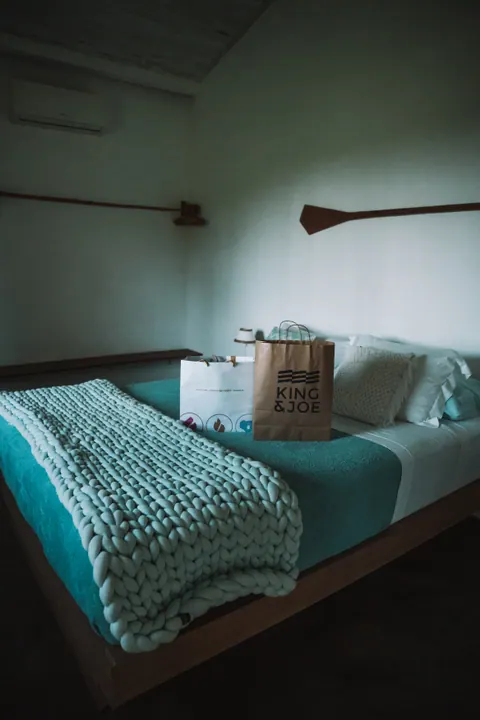 Cama com manta de tricô e duas sacolas de papel, uma delas da King & Joe, em um quarto, na primeira edição do Lanú.Lab, em Itacaré, na Bahia, em novembro de 2025.