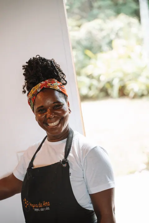 Mulher de avental preto e faixa estampada no cabelo sorri para a câmera, com vegetação ao fundo, na primeira edição do Lanú.Lab, em Itacaré, na Bahia, em novembro de 2025.