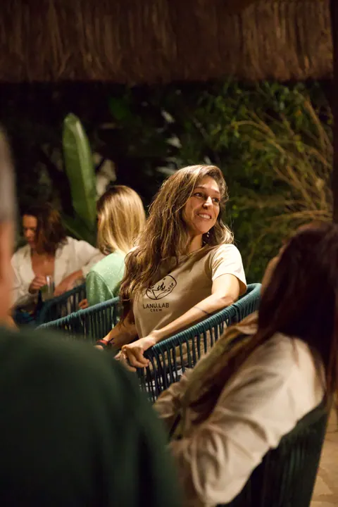Mulher com camiseta Lanú.Lab Crew sorri, sentada entre outras pessoas, à noite, na segunda edição do Lanú.Lab, na Chapada dos Veadeiros, em agosto de 2026.