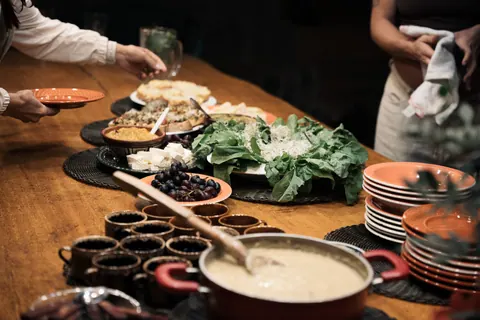 Mesa de madeira posta com saladas, queijos, uvas, canecas de cerâmica e uma panela, enquanto pessoas se servem, na segunda edição do Lanú.Lab, na Chapada dos Veadeiros, em agosto de 2026.