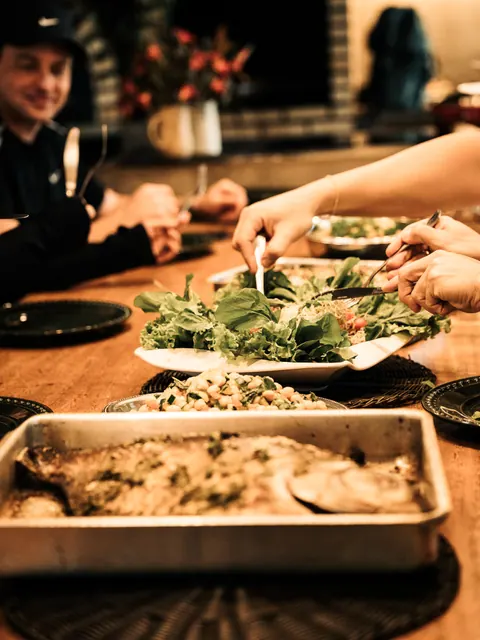 Mãos servem salada em uma mesa de madeira, com uma travessa de peixe assado em primeiro plano, na segunda edição do Lanú.Lab, na Chapada dos Veadeiros, em agosto de 2026.
