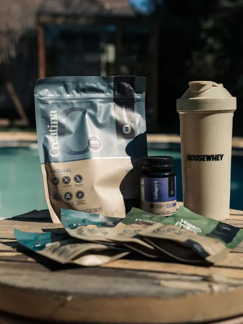 Embalagens de creatina, colágeno e proteína e uma coqueteleira da House Whey sobre uma mesa de madeira, com uma piscina ao fundo, na segunda edição do Lanú.Lab, na Chapada dos Veadeiros, em agosto de 2026.